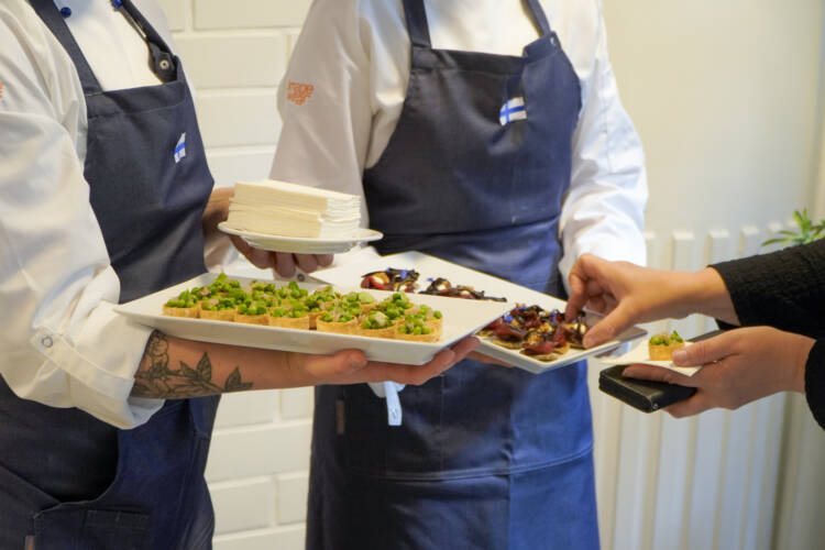 Kaksi kokinasuista henkilöä pitelemässä ruokatarjottimia, joissa on pieniä annospaloja. Kolmannen henkilön kädet ovat ottamassa ruokia.