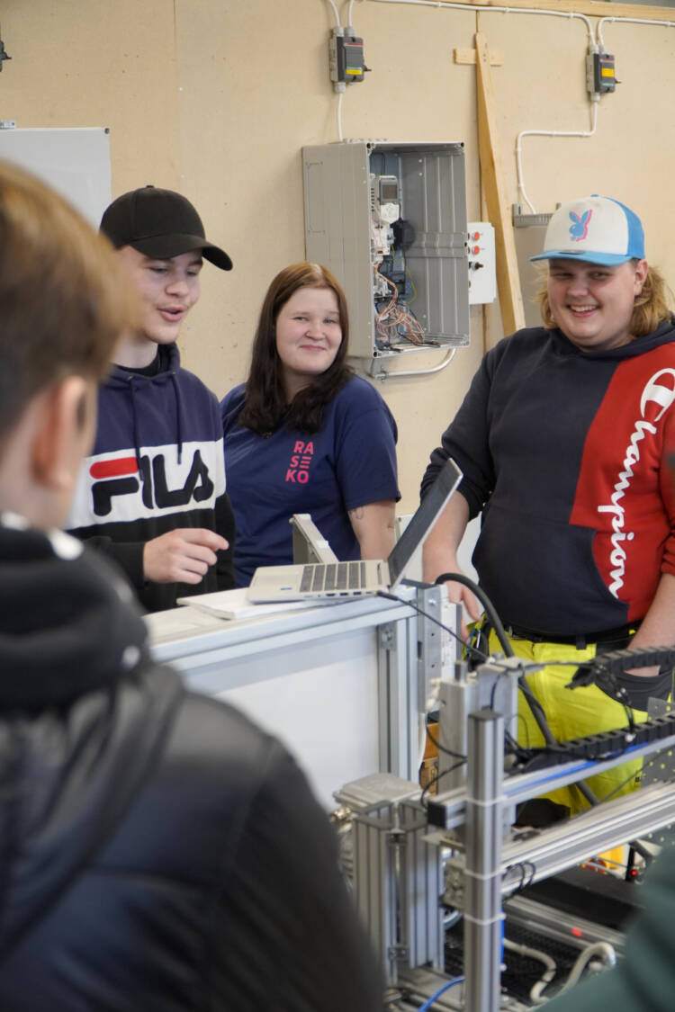 Kolme henkilöä seisoo ja hymyilee automaatiolaitteen takana.