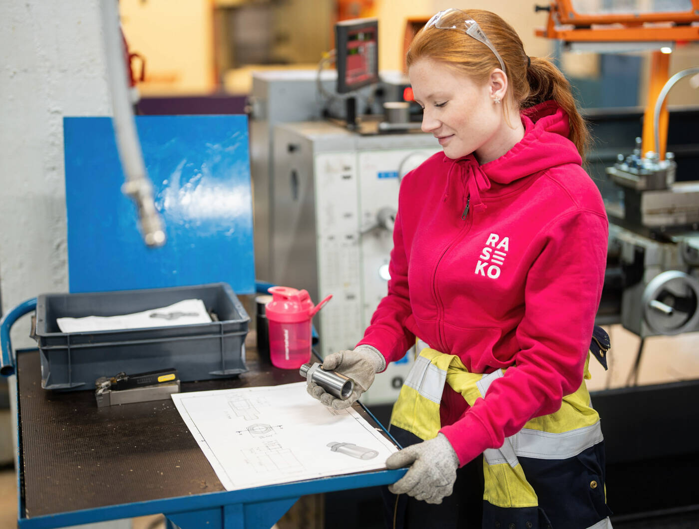 Pinkkiin Raseko-huppariin pukeutunut henkilö koneistamossa.