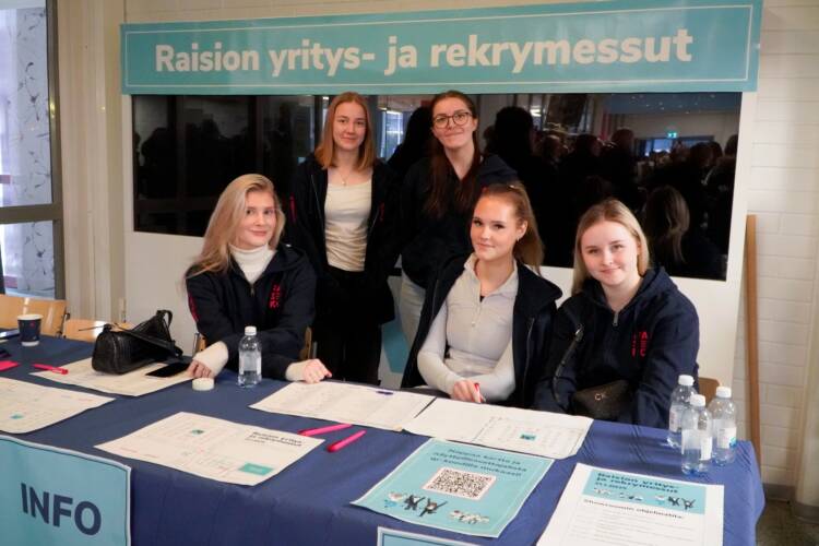 Viisi nuorta henkilöä hymyilee "Raision yritys- ja rekrymessut" -banderollin alapuolella.